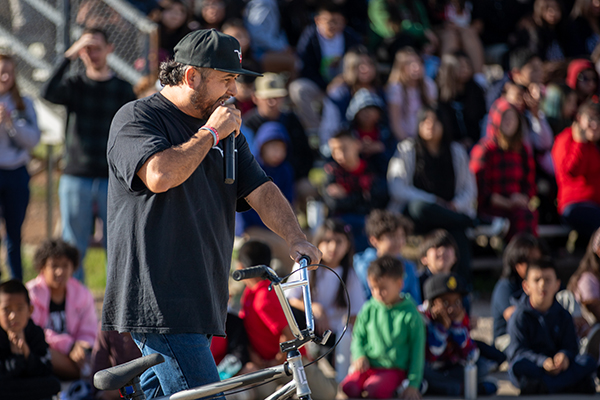 A man holding a BMX bike talks into a microphone to a crowd of students